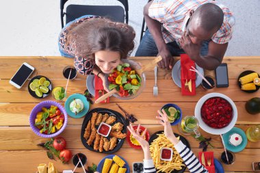 Ahşap masada otururken birlikte yemek yiyen bir grup insanın üst düzey görüntüsü. Masada yemek var. İnsanlar fast food yiyor..
