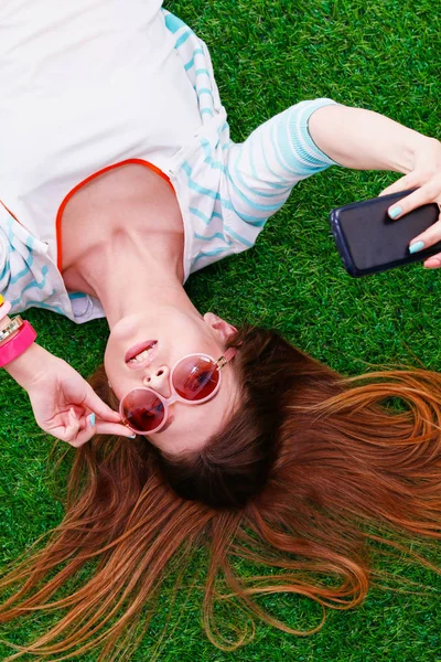 Mooie jonge vrouw die selfie maakt door haar telefoon terwijl ze in groen gras ligt. mooie jonge vrouw maken selfie — Stockfoto