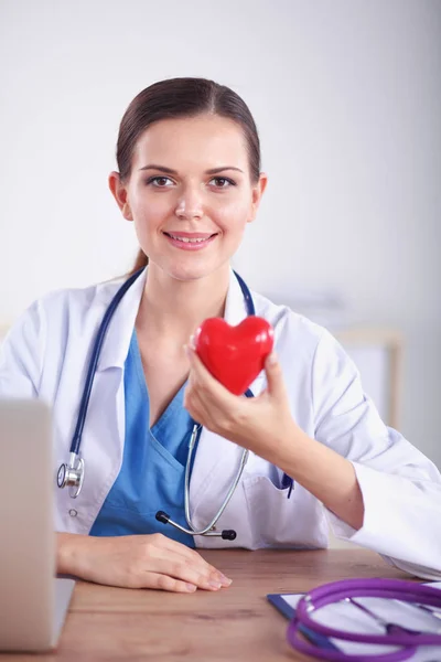 Schöne junge lächelnde Ärztin sitzt am Schreibtisch und hält Herz. — Stockfoto