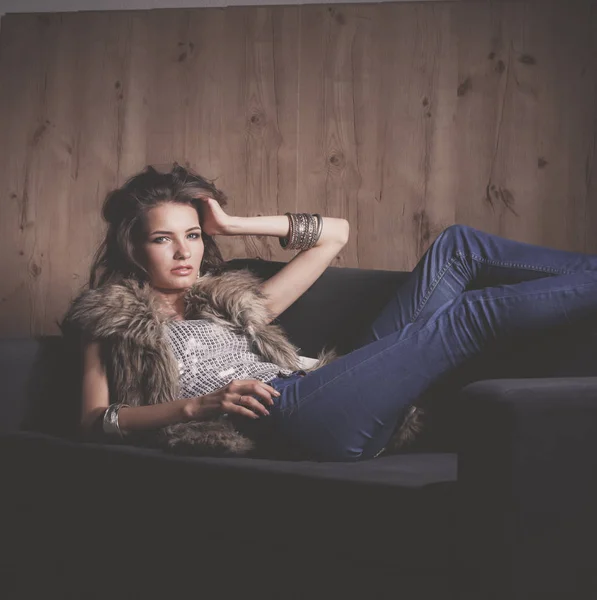Portrait of elegant woman sitting on black sofa wearing a blue jeans and fur vest — Stock Photo, Image