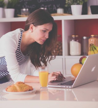 Dizüstü bilgisayar kullanarak ve mutfakta oturmuş çekici genç kadın