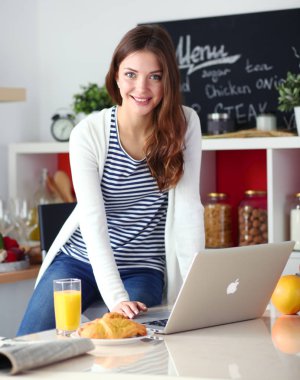 Dizüstü bilgisayar kullanarak ve mutfakta oturmuş çekici genç kadın