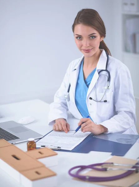 Piękna młoda uśmiechnięta lekarka siedzi przy biurku i pisze — Zdjęcie stockowe