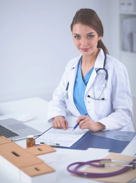 Hermosa joven sonriente doctora sentada en el escritorio y escribiendo —  Fotos de Stock