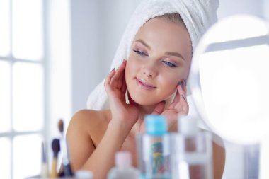 Bornozlu genç kadın banyo aynasına bakıyor.