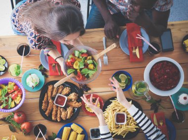 Ahşap masada otururken birlikte yemek yiyen bir grup insanın üst düzey görüntüsü. Masada yemek var. İnsanlar fast food yiyor..