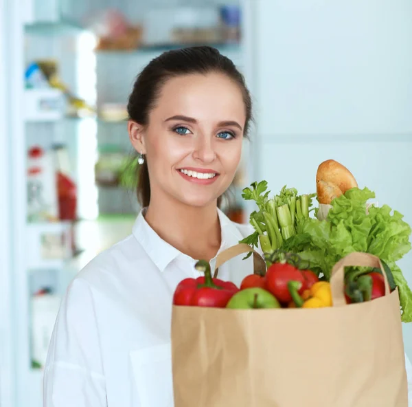 Fiatal nő gazdaság élelmiszer bevásárló táska zöldségekkel.Álló a konyhában — Stock Fotó