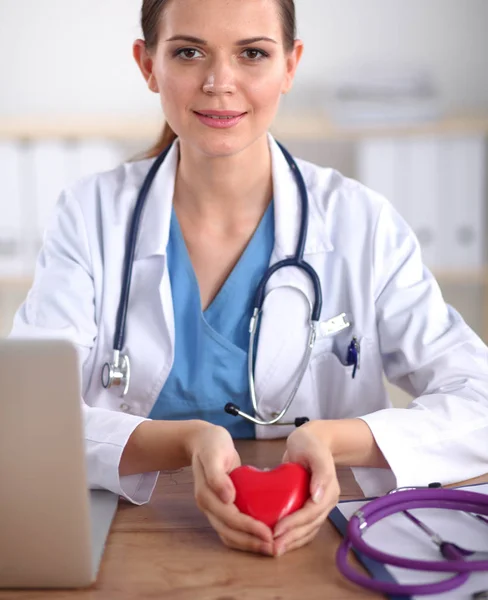 Young woman doctor holding a red heart sitting on the desk — Stock Photo, Image