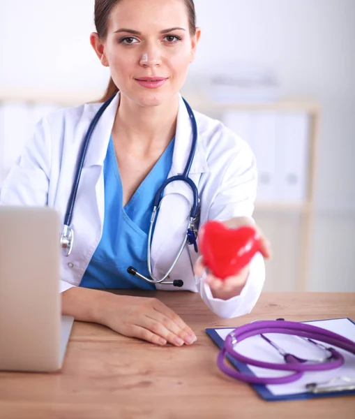 Schöne junge lächelnde Ärztin sitzt am Schreibtisch und hält Herz — Stockfoto