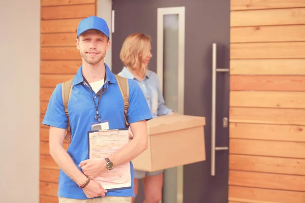 Lachende bezorger in blauw uniform levert pakketdoos aan ontvanger - koerier service concept. Glimlachende bezorger in blauw uniform — Stockfoto