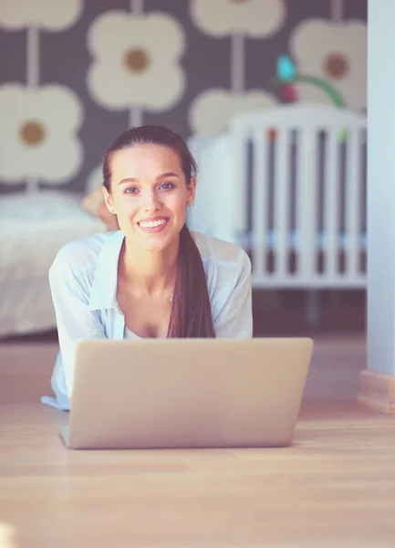 Ung kvinna som sitter på golvet nära barnens babysäng med laptop. Ung mamma — Stockfoto