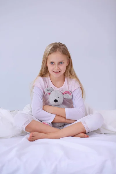 Liten flicka i sovrummet sitter på sängen. Lilla flickan bär en pyjamas och sitter i sängen . — Stockfoto