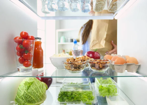 Portrait of female standing near open fridge full of healthy food, vegetables and fruits. — Stock Photo, Image