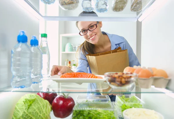 Porträt einer Frau, die neben einem offenen Kühlschrank voller gesunder Lebensmittel, Gemüse und Obst steht. — Stockfoto