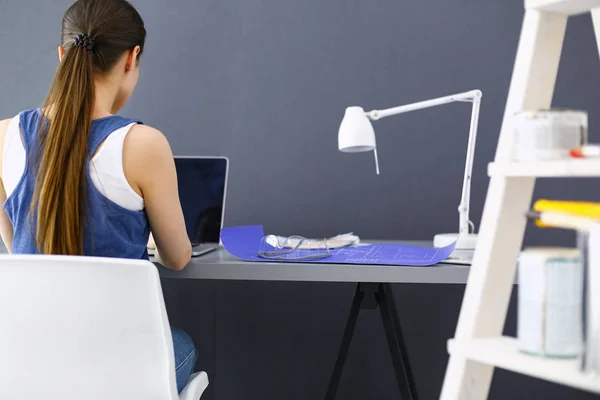 Ung kvinna sitter vid skrivbordet med instrument, plan och laptop. — Stockfoto