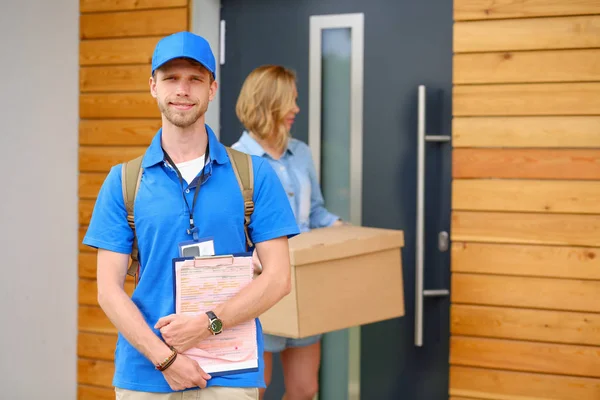 Repartidor sonriente con uniforme azul que entrega la caja de paquetes al destinatario: concepto de servicio de mensajería. Repartidor sonriente en uniforme azul —  Fotos de Stock