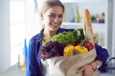 Sebzeli alışveriş poşeti taşıyan genç bir kadın. Mutfakta duruyor.