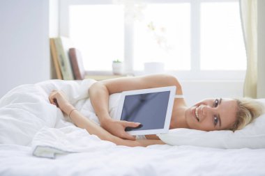 Elinde boş ekranlı dijital tablet tutan ve yatak odasında kameraya gülümseyen bir kız.