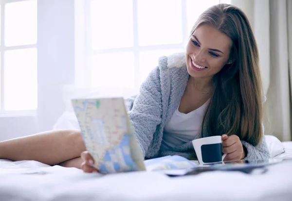 Avslappnad ung kvinna sitter på sängen med en kopp kaffe och digital tablett — Stockfoto