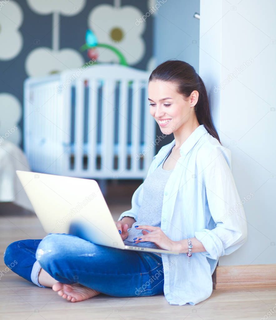 Young woman sitting on the floor near childrens cot with laptop. Young mom