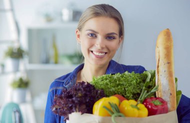 Sebzeli alışveriş poşeti taşıyan genç bir kadın. Mutfakta duruyor.