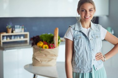 Sebzeli alışveriş poşeti taşıyan genç bir kadın. Mutfakta duruyor.