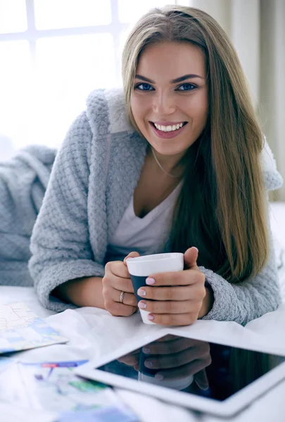 Giovane donna rilassata seduta sul letto con una tazza di caffè e tablet digitale — Foto Stock