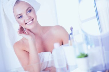 Bornozlu genç kadın banyo aynasına bakıyor.