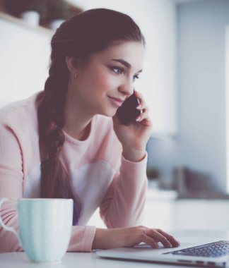 Genç kadın bir dizüstü bilgisayar kullanarak ve cep telefonuyla konuşurken mutfak masasında oturur.