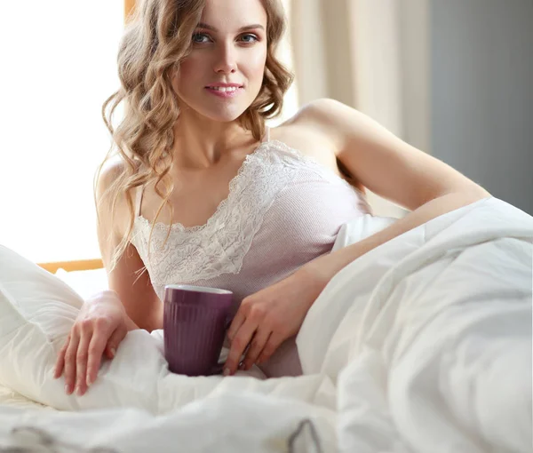 Mujer joven bebiendo taza de café o té mientras está acostada en la cama . — Foto de Stock