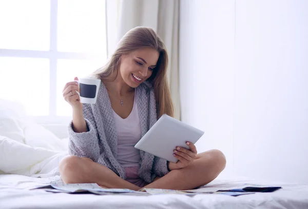 Bir fincan kahve ve dijital tabletle yatakta oturan rahat genç bir kadın. — Stok fotoğraf