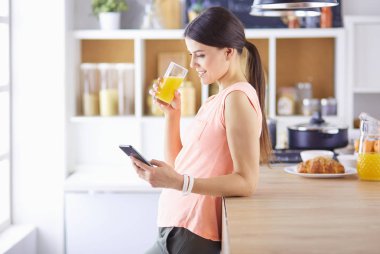 Cep telefonu görünümlü ve cam portakal suyu bir mutfakta Kahvaltı yaparken tutan gülümseyen güzel kadın.