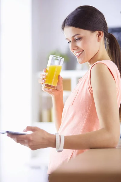 Усміхнена красива жінка дивиться на мобільний телефон і тримає склянку апельсинового соку під час сніданку на кухні . — стокове фото