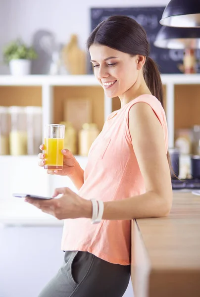 Leende vackra kvinna tittar på mobiltelefon och hålla glas apelsinjuice medan du äter frukost i ett kök. — Stockfoto