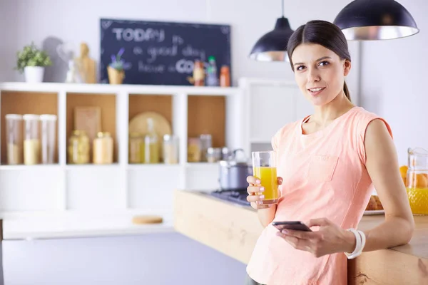 Mujer bonita sonriente mirando el teléfono móvil y sosteniendo un vaso de jugo de naranja mientras desayunaba en una cocina . — Foto de Stock