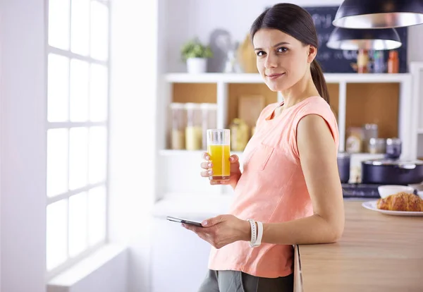 Leende vackra kvinna tittar på mobiltelefon och hålla glas apelsinjuice medan du äter frukost i ett kök. — Stockfoto