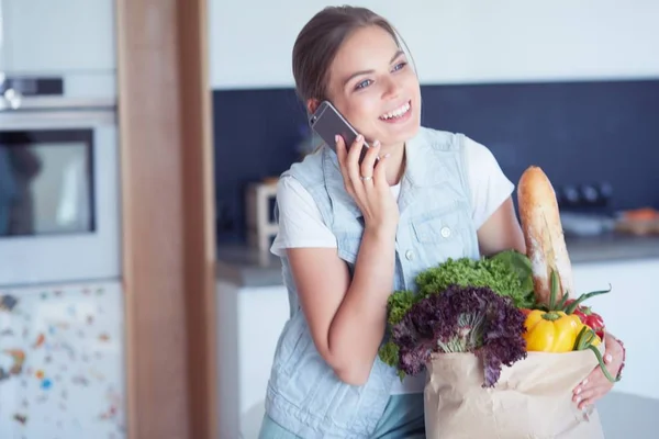 Mosolygós nő mobiltelefon gazdaság bevásárló táska a konyhában — Stock Fotó