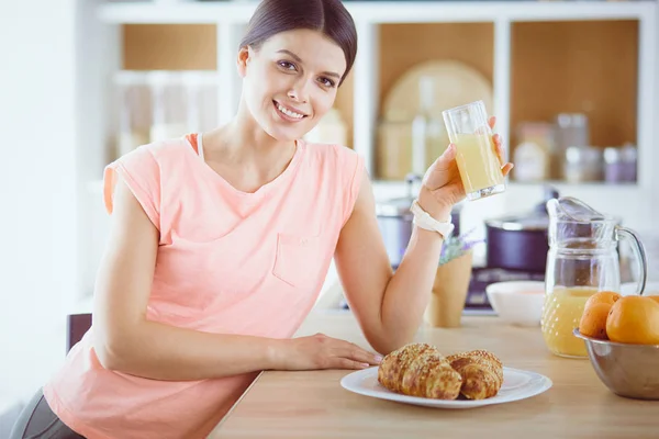 Leende vackra kvinna tittar på mobiltelefon och hålla glas apelsinjuice medan du äter frukost i ett kök. — Stockfoto