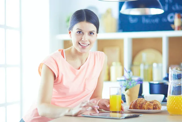 Leende vackra kvinna tittar på mobiltelefon och hålla glas apelsinjuice medan du äter frukost i ett kök. — Stockfoto