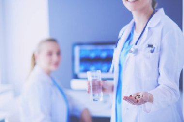 Doktorun arka planda meslektaşıyla klinikte elinde haplar ve bir bardak su tutarken görüntüsü..