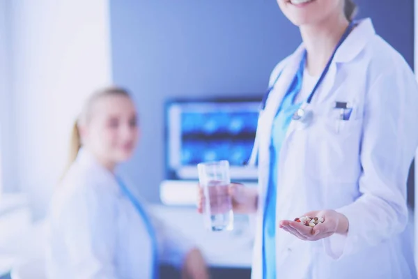 Inyección del médico sosteniendo pastillas y un vaso de agua en la clínica con su colega en el fondo. —  Fotos de Stock