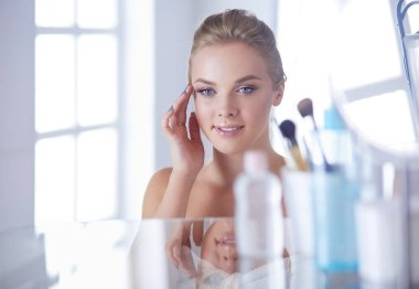 Bornozlu genç kadın banyo aynasına bakıyor.