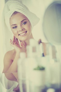 Bornozlu genç kadın banyo aynasına bakıyor.