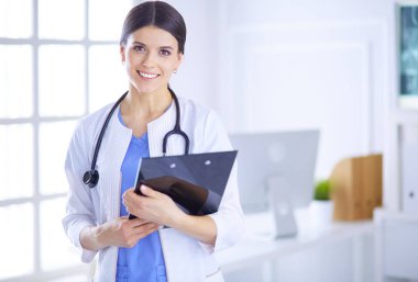 Steteskoplu gülümseyen genç bayan doktor hastane danışma odasında bir dosya tutuyor.