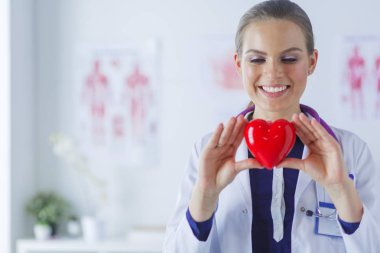 Steteskoplu bir doktor, beyaz arka planda kırmızı kalbi inceliyor.