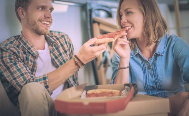 Genç çift, yeni bir eve taşındıktan sonra etraflarında kutularla yerde pizza molası veriyorlar. Genç çift