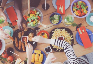 Ahşap masada otururken birlikte yemek yiyen bir grup insanın üst düzey görüntüsü. Masada yemek var. İnsanlar fast food yiyor..