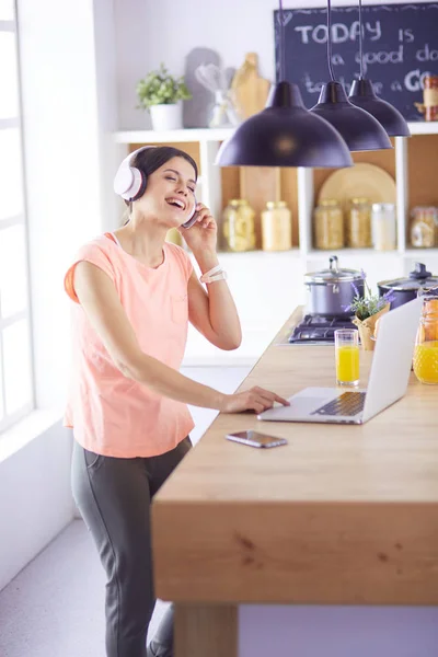 Portret van een vrolijke jonge vrouw die naar muziek luistert met een koptelefoon en met laptop computer terwijl hij in de keuken staat — Stockfoto