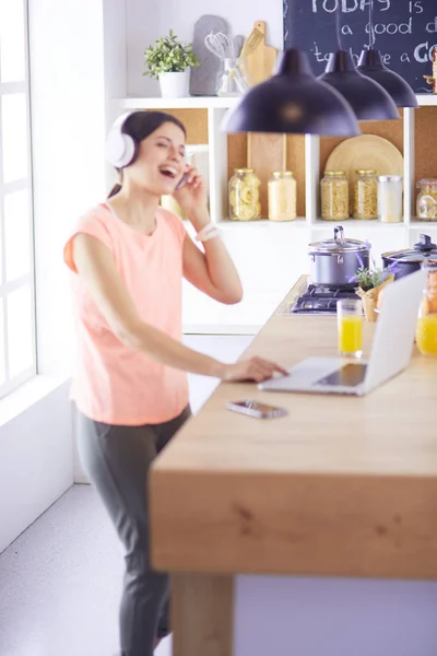 Portrét veselé mladé ženy, která poslouchala hudbu se sluchátky a používá přenosný počítač, zatímco stojí v kuchyni — Stock fotografie