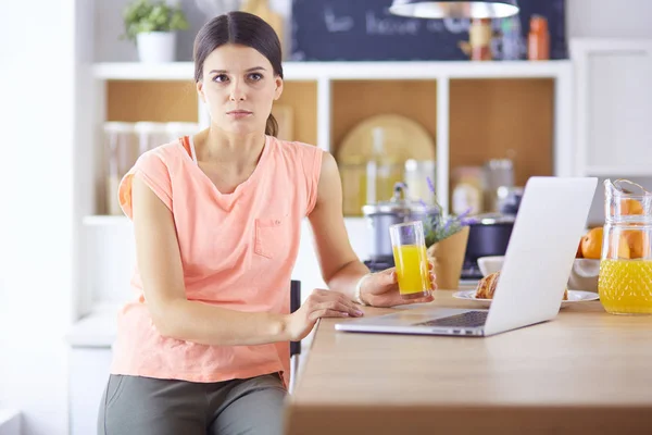 Mujer joven en la cocina con computadora portátil buscando recetas, sonriendo. Concepto de bloguero de alimentos — Foto de Stock